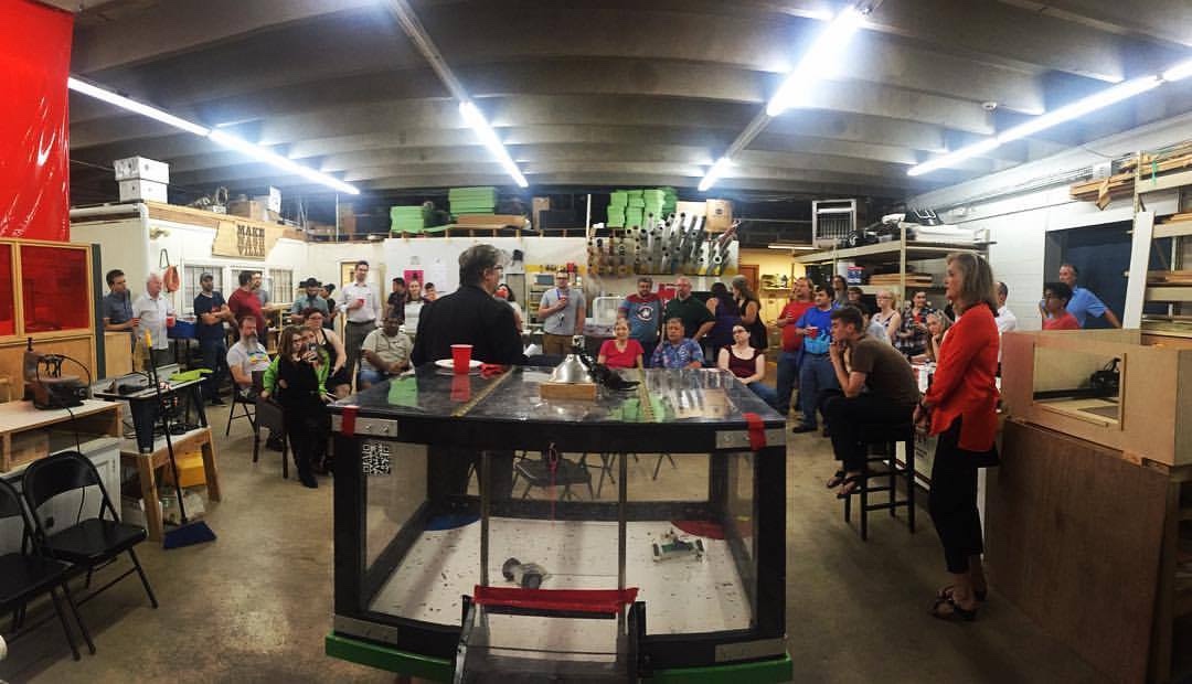 A crowd of members gathered around a clear acrylic combat-robot arena at the Woodland Street space, with the Tennessee-shaped MAKE NASHVILLE logo on the wall behind them.